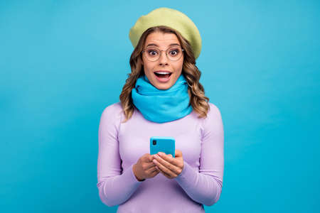 Portrait of astonished excited crazy girl use smartphone impressed social network notification scream wow omg wear trendy violet jumper green headwear isolated over blue color backgroundの写真素材