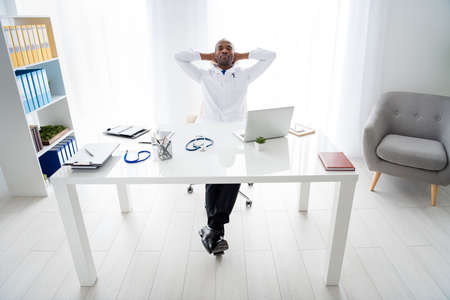 Full length photo of family doc dark skin guy help control epidemic disease patients working day night need some rest hold hands behind head wear white lab coat office clinic indoorsの写真素材