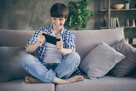 Photo of teenager school boy enjoy relaxing sit comfy couch barefoot legs crossed stay home quarantine time hands hold telephone addicted gamer play online living room indoorsの写真素材