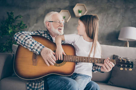 Photo of aged old grandpa little pretty granddaughter sitting comfy sofa playing guitar teaching bonding spend time together stay home quarantine safety modern interior living room indoorsの写真素材