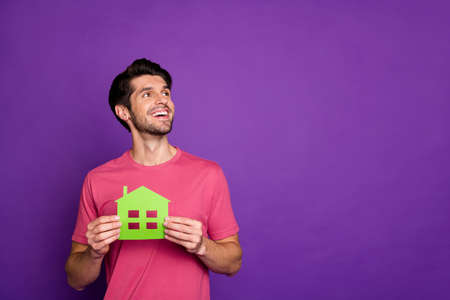 Photo of attractive handsome guy good mood hold green paper house building hands real estate agent look empty space wear casual pink t-shirt isolated purple color backgroundの写真素材