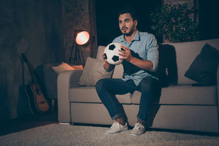 Portrait of his he nice attractive addicted nervous brunet guy sitting on divan watching sport show holding in hands soccer ball at modern industrial loft style interior dark roomの写真素材