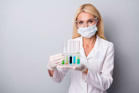 Portrait of expert retired pensioner woman hold flask with chemical liquid corona virus infection antidote vaccination wear mask goggles gloves rubber isolated gray color backgroundの写真素材