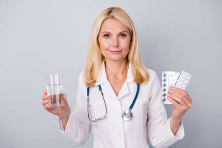 Portrait of specialist woman doctor hold pills glass water wear stethoscope isolated over gray color backgroundの写真素材