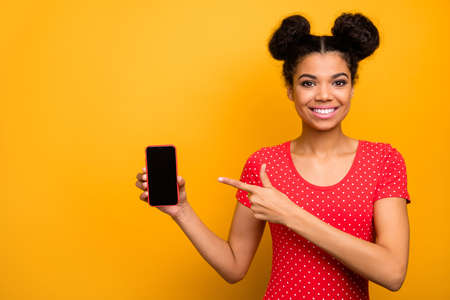 Portrait of positive cheerful afro american girl promoter show smartphone point index finger present new modern device wear red dotted t-shirt isolated over bright shine color backgroundの写真素材