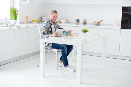 Full length photo  positive guy have brunch eat sweet tasty delicious muesli granola use tablet watch funny  blog video wear casual plaid denim sit kitchen table in house indoorsの写真素材