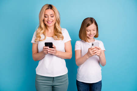 Photo of two people beautiful mom lady little daughter blonds spend time together hold telephones hands writing new post blog wear casual white s-shirts isolated blue color backgroundの写真素材