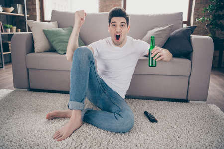 Portrait of his he nice attractive addicted cheerful cheery guy sitting on carpet spending day watching TV show drinking beer having fun at modern industrial loft brick interior style living-roomの写真素材