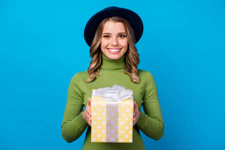 Photo of attractive cheerful lady hold big yellow dotted gift box birthday present party guest wear retro hat casual green turtleneck isolated blue color backgroundの写真素材