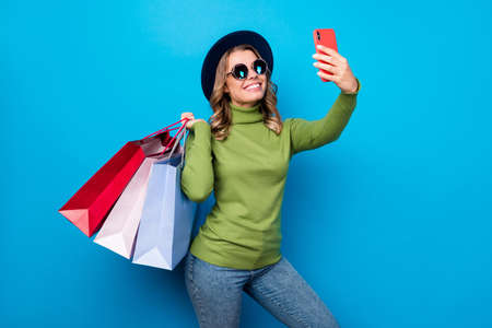 Photo of cheerful funny curly lady hold many shop packs shopaholic making selfies telephone popular blogger wear sun specs hat casual green turtleneck jeans isolated blue color backgroundの写真素材