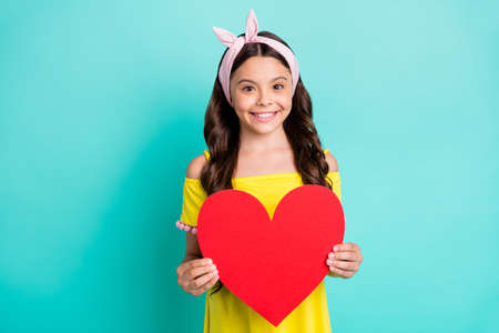 Portrait of positive girl kid hold big red paper card heart enjoy dreamy romantic date 14-february present event wear stylish trendy clothes isolated over turquoise color backgroundの写真素材