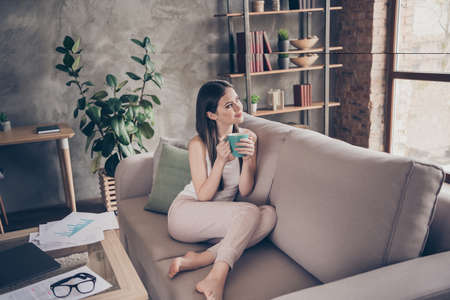 Full length photo of interested positive girl have free time after routine remote work hold beverage look dream dreamy sit divan in house indoorsの写真素材
