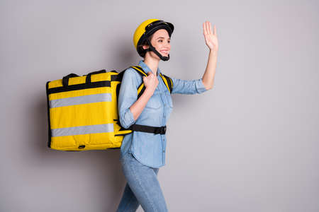 Profile side photo positive girl courier go walk deliver customer client meal look copyspace see friend greet raise palm wear denim shirt helmet thermal bag isolated gray color backgroundの写真素材