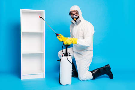 Full body photo of man wear bio hazmat suit breathing mask use sprayer equipment disinfect shelves ncov infection isolated over blue color backgroundの写真素材