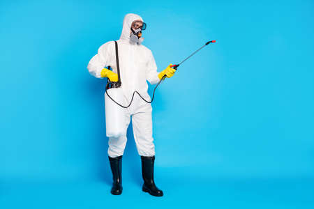 Full length photo of medical worker man use sprayer equipment disinfect copyspace covid infection spread wear latex gloves biohazard suit boots isolated over blue color backgroundの写真素材
