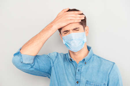 Portrait of ill sick young man in blue shirt wear medical safety face mask, touching his head...