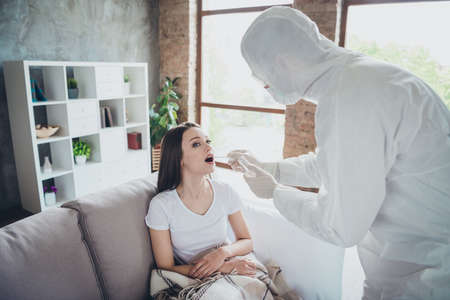 Photo of young patient lady alone sitting sofa feel unwell ambulance emergency service guy doc virology clinic collect saliva mouth probe wear uniform protection visit her flat indoorsの写真素材