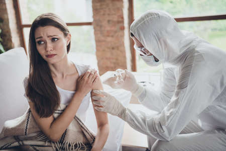 Photo of young sick patient lady sit sofa feel unwell call emergency doc virologist flu cold covid injection hand shoulder medication help wear latex gloves protective uniform indoorsの写真素材