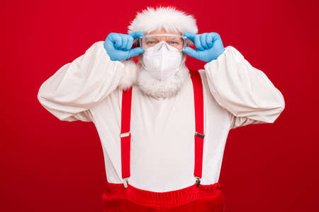 Photo of old merry cheerful santa working every day dont mind epidemic restrictions x-mas spirits soul wear gloves goggles face mask hat shirt suspenders isolated red backgroundの写真素材