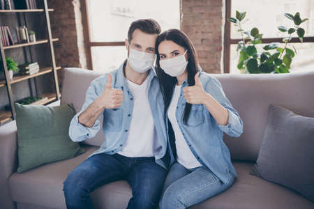 Portrait of positive two people married couple stay home want cov infection epidemic protection wear medical mask recommend show thumb up sign in house indoorsの写真素材