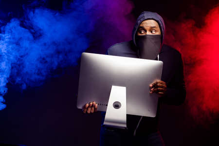 Photo of worried scared afro american guy rob computer pc screen, look copyspace check police dont chase wear hood sweater isolated black fog color backgroundの写真素材