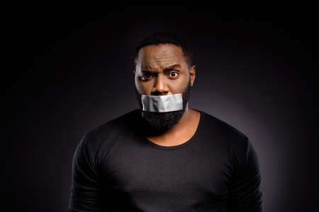 Close up photo of shocked afro american guy close cover lips mouth adhesive tape speechless voiceless african community member racism indifference isolated over black color backgroundの写真素材