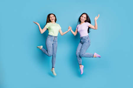 Full length photo of two people lesbians couple ladies jump high up hold hands good mood rejoicing happy together wear casual t-shirts jeans footwear isolated blue color backgroundの写真素材