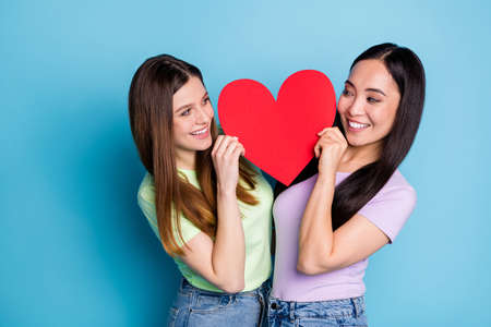Photo of two funny lesbians couple ladies romance date hold big red paper heart shape lovers look eyes tricky flirty cunning wear casual t-shirts jeans isolated blue color backgroundの写真素材