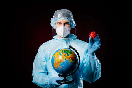 Photo of serious guy doc virologist found vaccine hold globe allow patients travel wear respirator hazmat uniform suit plastic facial protective shield isolated black color backgroundの写真素材