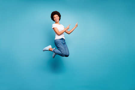 Full body photo of funny dark skin wavy lady jumping high directing fingers empty space offer black friday prices wear casual white t-shirt jeans isolated blue color backgroundの写真素材