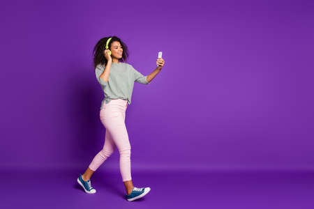Full size photo of positive afro american girl break pause go walk listen music choose song cellphone have ear phones wear striped shirt pink trousers sneakers isolated purple color backgroundの写真素材