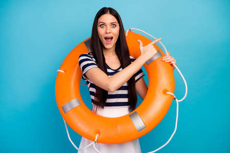 Photo of excited lady long hair hold orange emergency life buoy directing finger empty space advising cheap cruise liner trip wear white striped summer dress isolated blue backgroundの写真素材