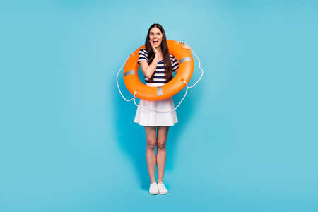Full size photo of amazed lady crazy tourist long pretty legs hold orange emergency life buoy hand on cheek wear white striped sun short summer dress shoes isolated blue backgroundの写真素材