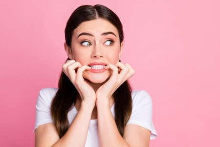 Closeup photo of pretty terrified lady long hairdo tails look side empty space eyes full of fear biting fingers panic nervous wear casual white t-shirt isolated pastel pink color backgroundの写真素材