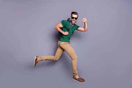 Full length profile side photo of excited guy jump run enjoy summer weekend tour hold backpack wear green t-shirt pants trousers isolated over gray color backgroundの写真素材