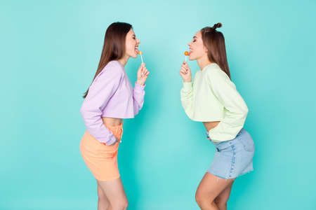 Profile photo of two funny ladies standing opposite hold lollipop chupa chups hands childish mood tasty sweets wear cropped pullovers mini skirts isolated pastel teal color backgroundの写真素材