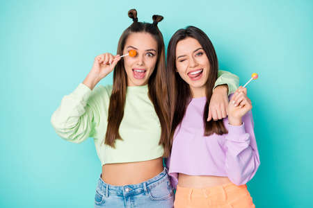Photo of two funny cheerful ladies hold lollipop chupa chups hands crazy childish mood playful wear cropped pullovers naked belly skirts isolated pastel teal color backgroundの写真素材