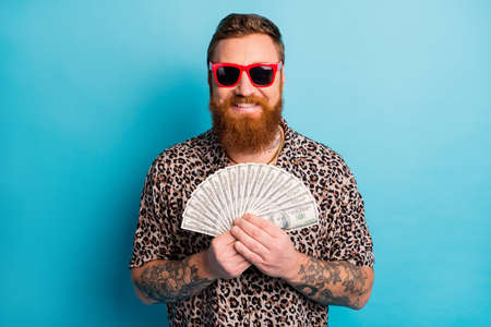 Photo of cool macho guy traveler holding fan of usa bucks excited good mood tattoo body hands ready spend waste money wear leopard shirt sun specs isolated blue backgroundの写真素材