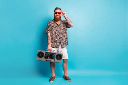 Full length photo of macho guy holding retro tape recorder modern youth party good mood ready chill rest rejoice wear shorts leopard shirt shoes sun specs isolated blue backgroundの写真素材