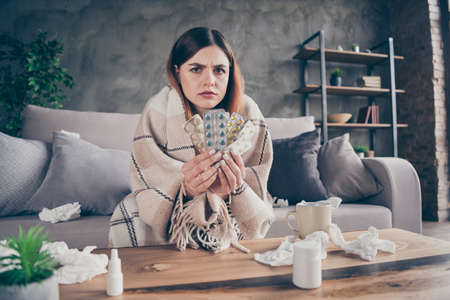 Portrait of frustrated ill girl sit divan suffer corona virus infection symptom hold pills tablets doctor physician pharmacist prescribe her cover checkered plaid blanket in house indoorsの写真素材