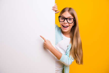 Photo of beautiful pretty little lady diligent student pupil holding hands direct finger big empty space white placard banner sales wear specs casual outfit isolated yellow color backgroundの写真素材