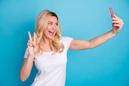 Photo of positive cheerful woman blogger enjoy weekend holiday photographing selfie smartphone make v-sign wear stylish clothes over blue color backgroundの写真素材