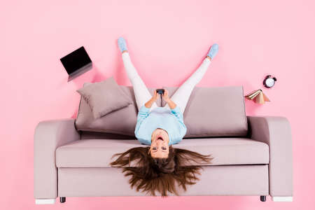 cheery girl sitting on divan upside down having fun using device isolated on pink pastel color backgroundの写真素材