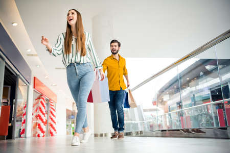 Low angle view full length photo of energetic woman see incredible 50 sales go walk hurry her husband follow hold many packages wear striped yellow shirt denim jeans in shopping mall centerの写真素材