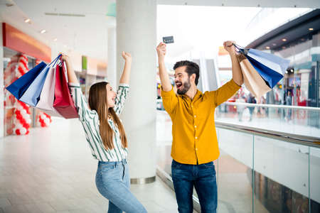 Photo of two people cheerful pretty lady handsome guy couple enjoy free time hold many bags walk shopping center raise hands use credit card discounts wear casual jeans shirt outfit indoorsの写真素材