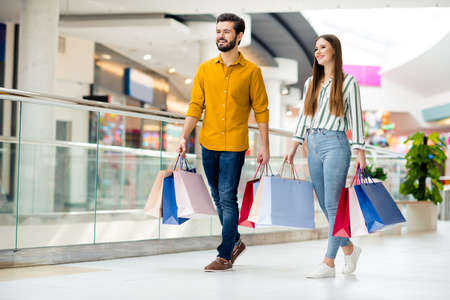 Full length photo of two people cheerful pretty lady handsome guy couple enjoy free time buy hold many bags walk shopping center floor wear casual jeans shirt shoes outfit indoorsの写真素材