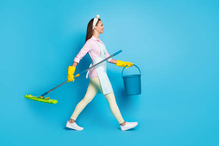 Full size profile side photo of positive professional cleaner worker girl hold mop bucket ready disinfect house covid infection wear pink shirt pants trousers isolated blue color backgroundの写真素材