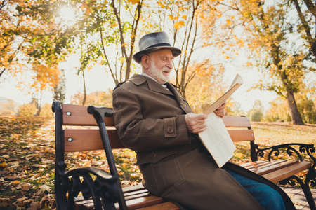 Profile side photo of old man have morning retired walk sit fall park bench outside read town center news wear long coat cap headwearの写真素材