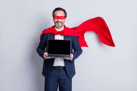 Photo of confident cheerful aged mature business guy super hero costume hold laptop presenting new device model sale price manager wear suit red face mask cloak isolated grey backgroundの写真素材