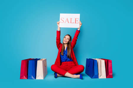 Full body photo of self-confident lady boss sitting floor shoes shopping bags raise sale placard wear red suit blouse shirt blazer trousers high-heels isolated blue color backgroundの写真素材